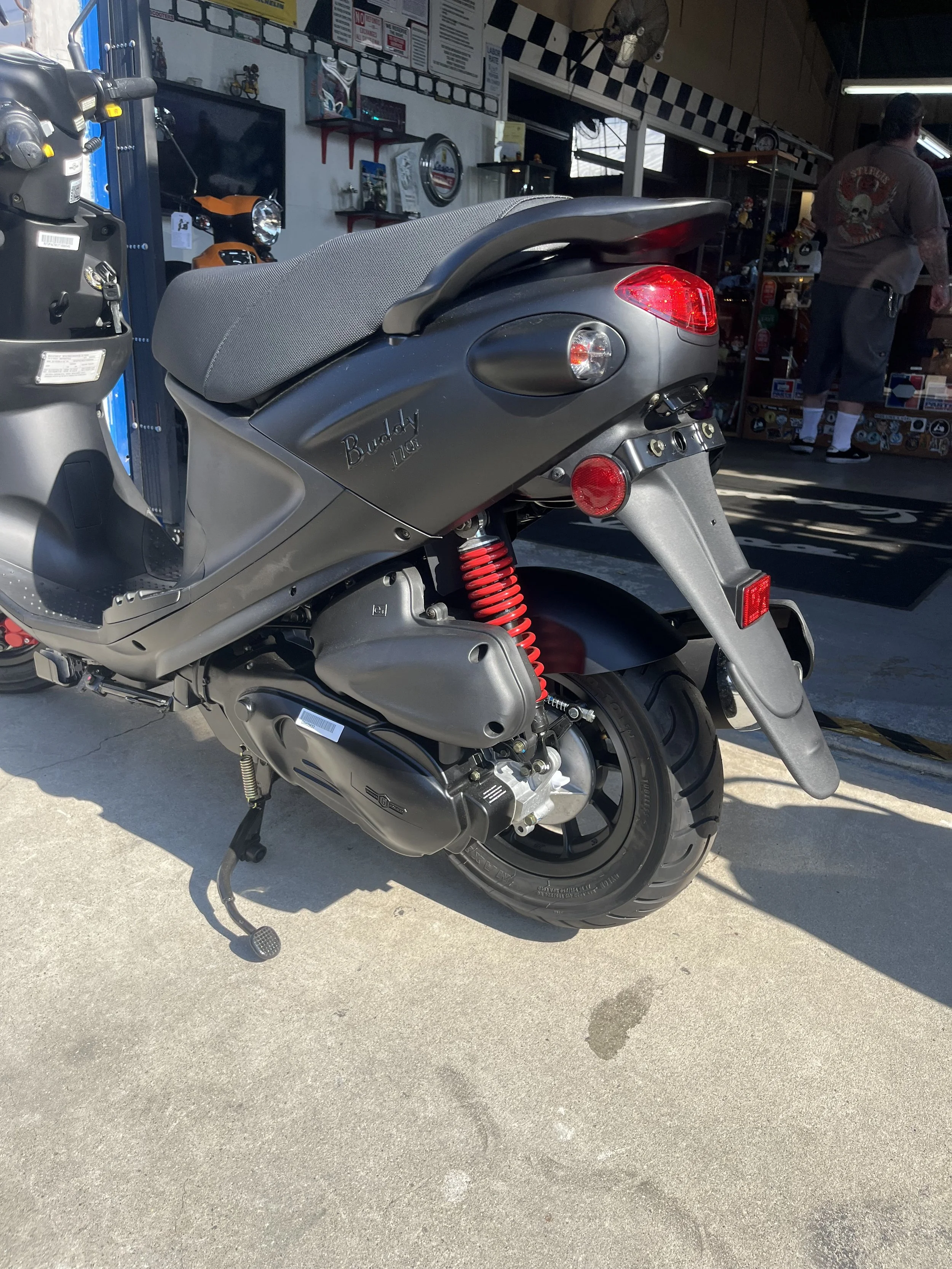 A matte gray Buddy scooter parked outside a store, showing its rear side with a red spring suspension, black wheel, and black exhaust. The store has a checkered pattern on the wall and some people inside.