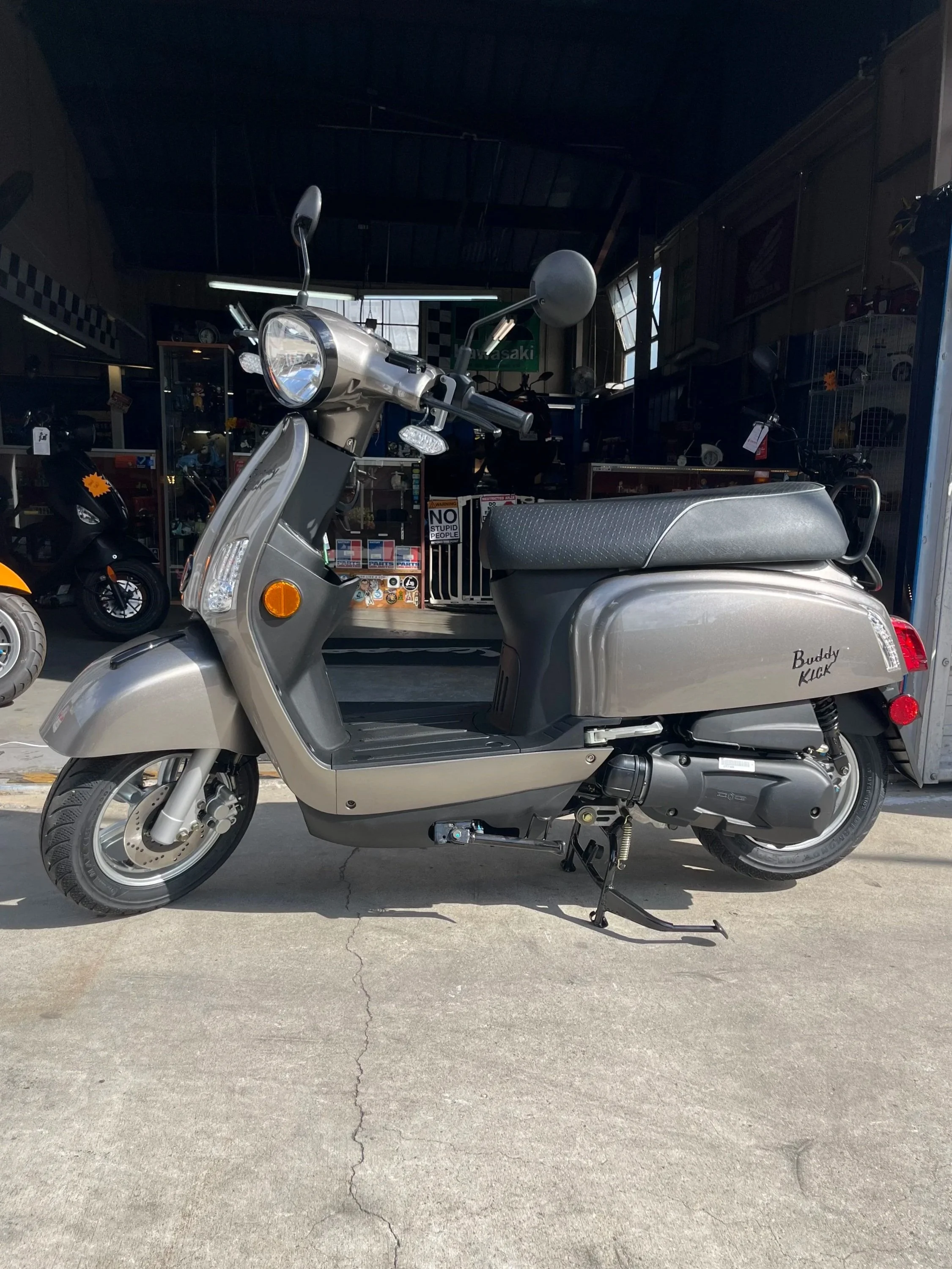 Gray Buddy Kick scooter parked on concrete outside a motorcycle shop.