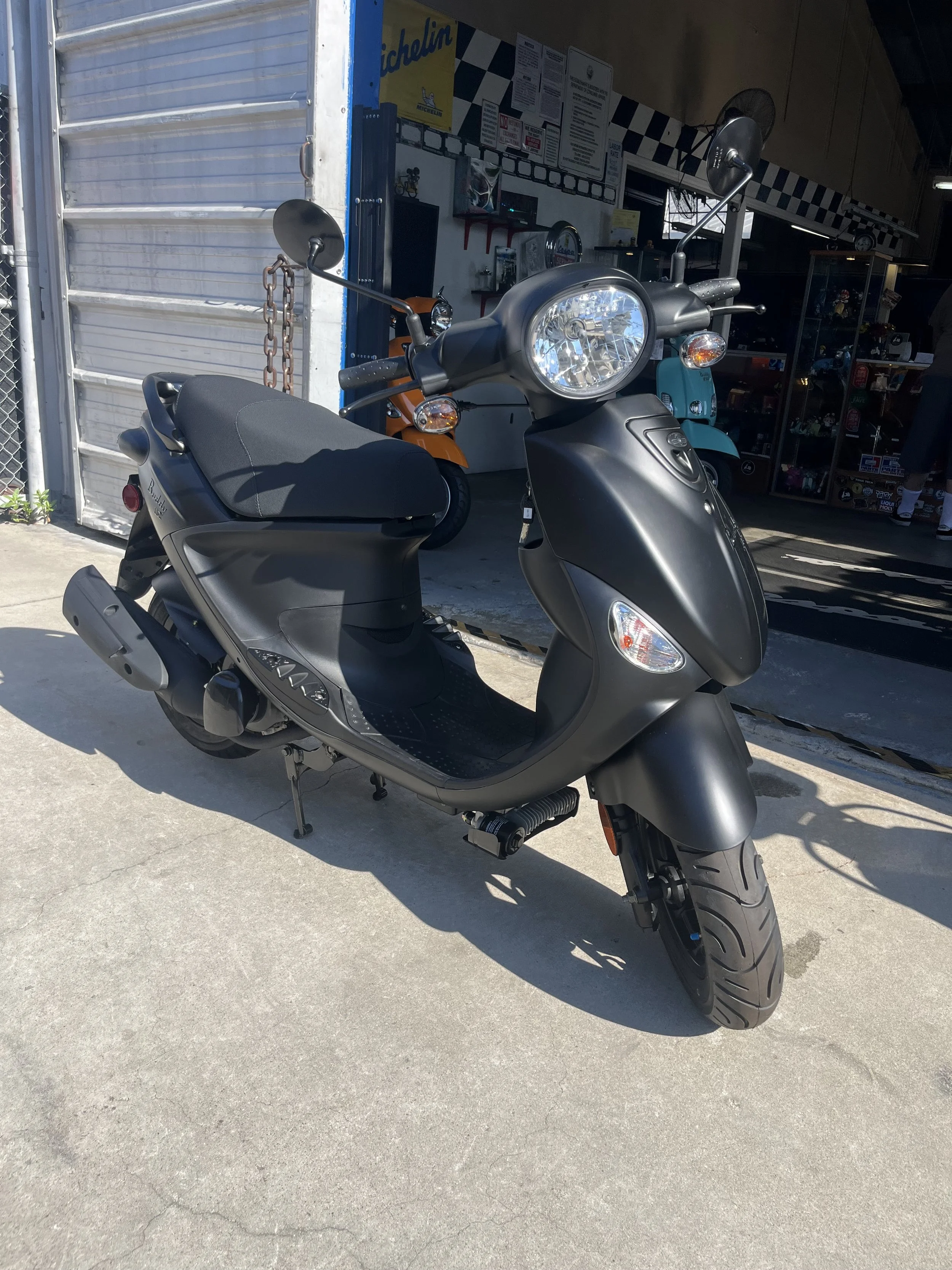 Black scooter parked outside a shop with motorcycles and a display case visible in the background.