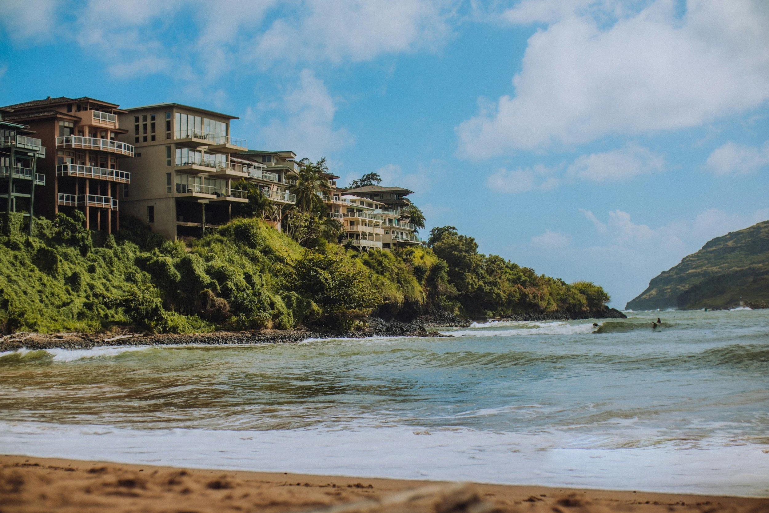 Beach with waves, green hills, and modern houses on the hillside under a blue sky with scattered clouds.