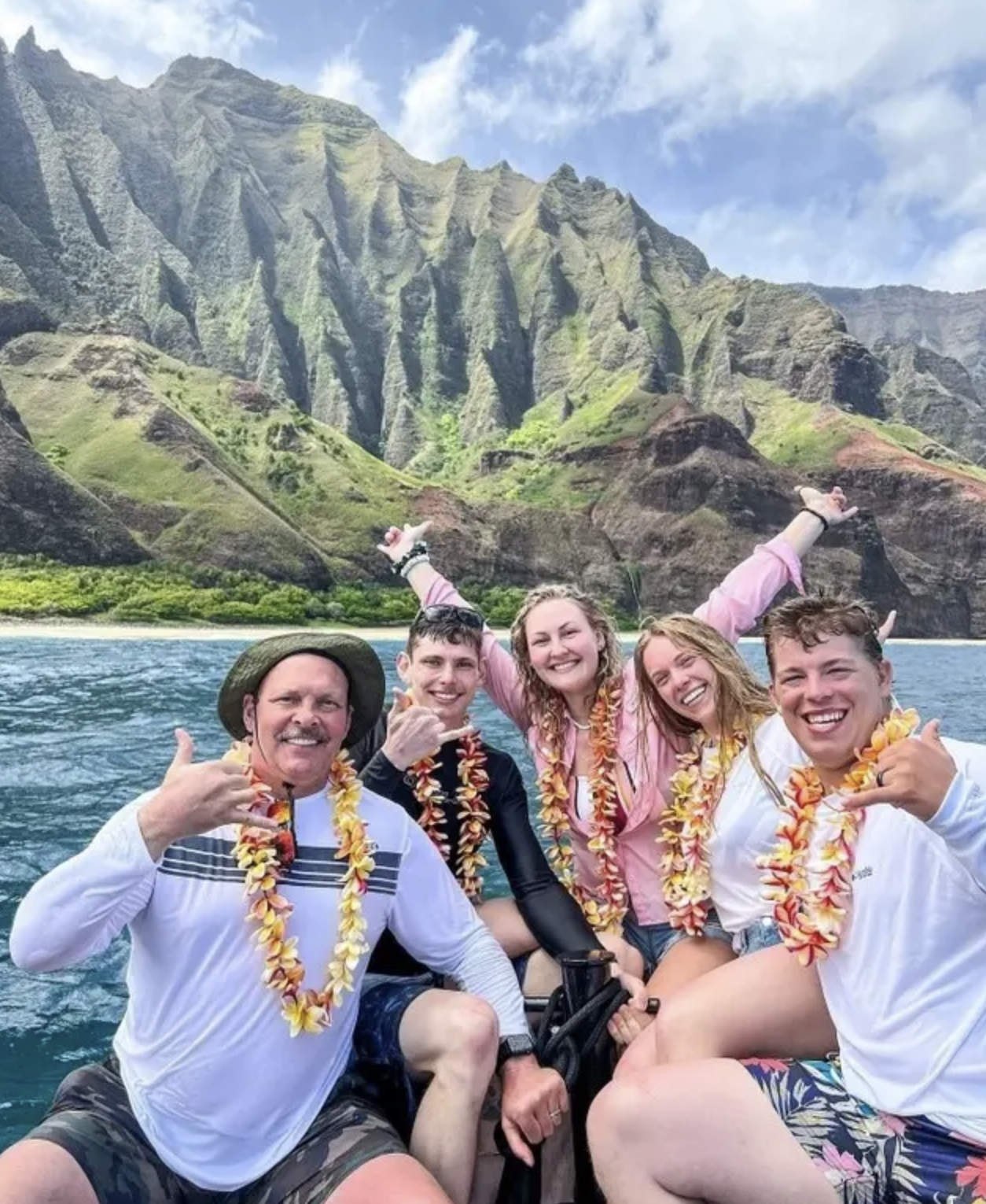 Group of five friends on a boat with a mountainous landscape in the background, wearing leis and smiling for the camera.