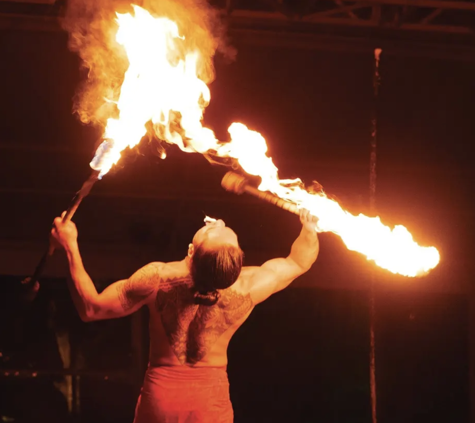 A fire performer is holding two flaming sticks and blowing fire in an outdoor setting at night.
