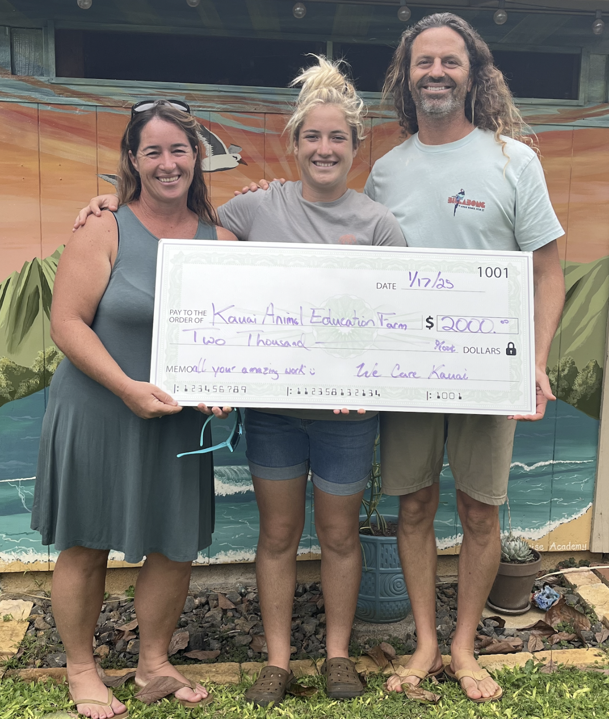 Three people standing outdoors holding a large check made out to the Kauai Animal Education Farm for $2000, smiling for the camera.