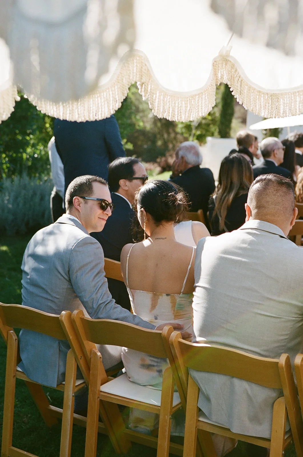 People attending outdoor wedding or event, sitting on wooden chairs, with a decorative canopy overhead and sunny weather.