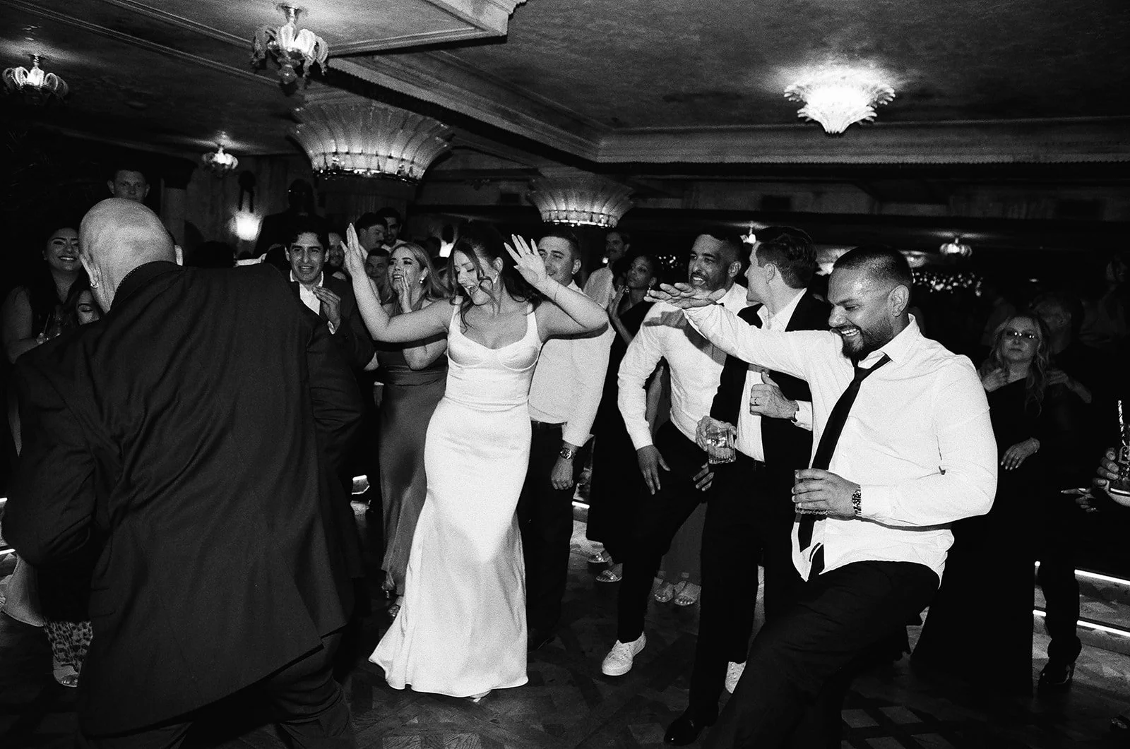 People dancing and enjoying themselves at a party or celebration, with some holding drinks, in an indoor venue with ornate ceiling lighting.