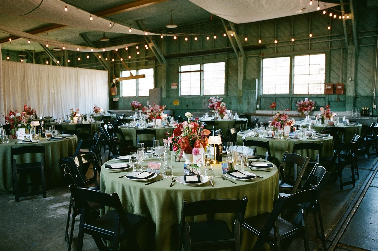 A large banquet hall decorated for an event with multiple round tables covered in green tablecloths. Each table has a centerpiece with pink and red flowers, glassware, and place settings. String lights hang from the ceiling, and the hall has green pa