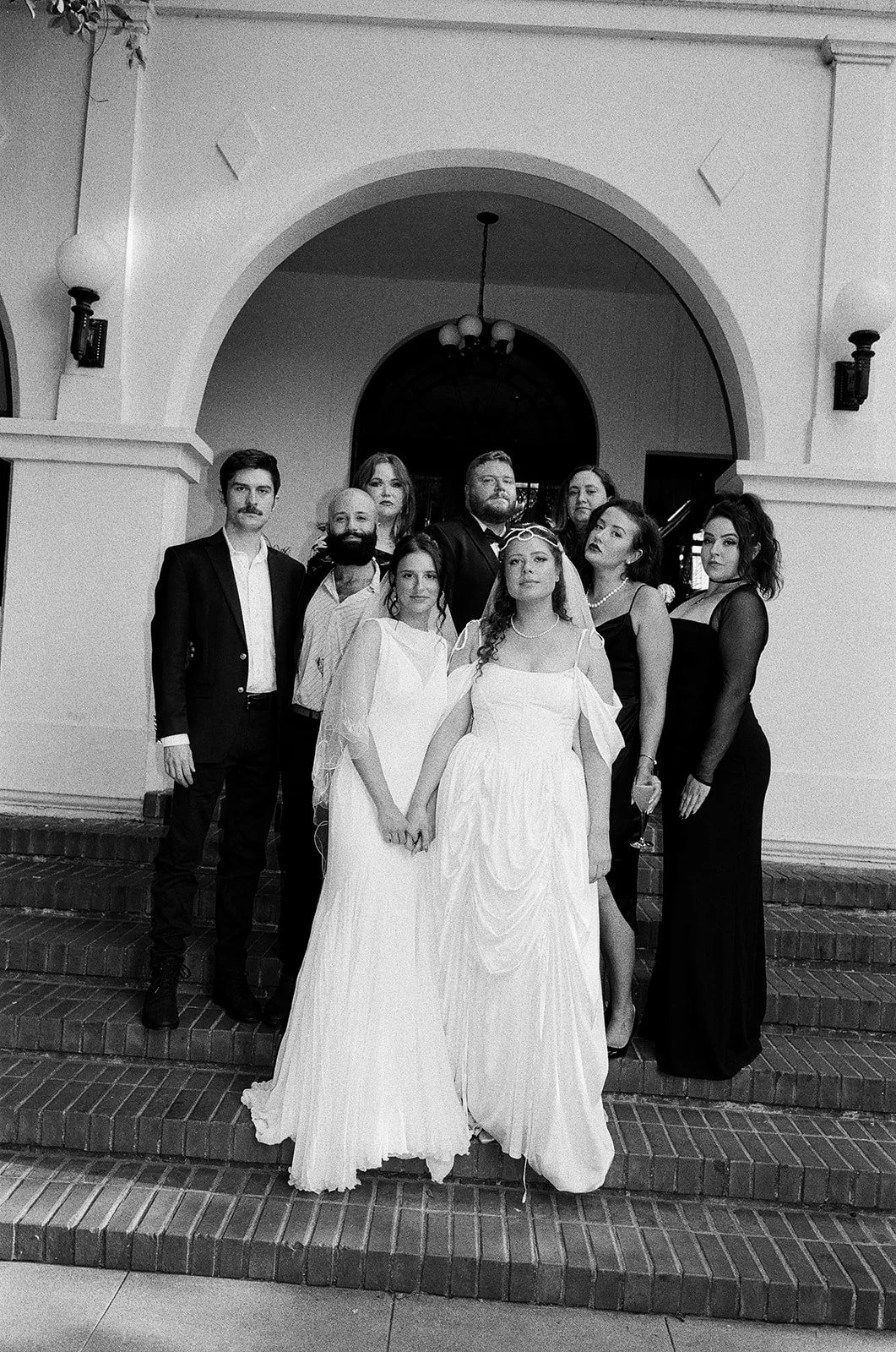 A group of people dressed in formal attire standing on steps in front of a building entrance, including two women in white gowns, indicating a wedding celebration.