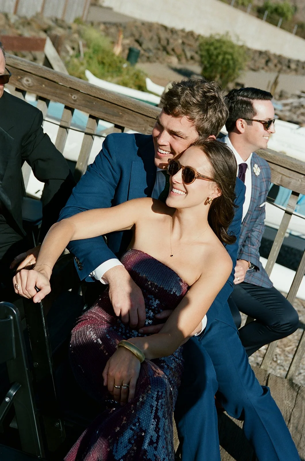 Young couple smiling and sitting close together on a dock, with other people in the background at a waterfront setting.