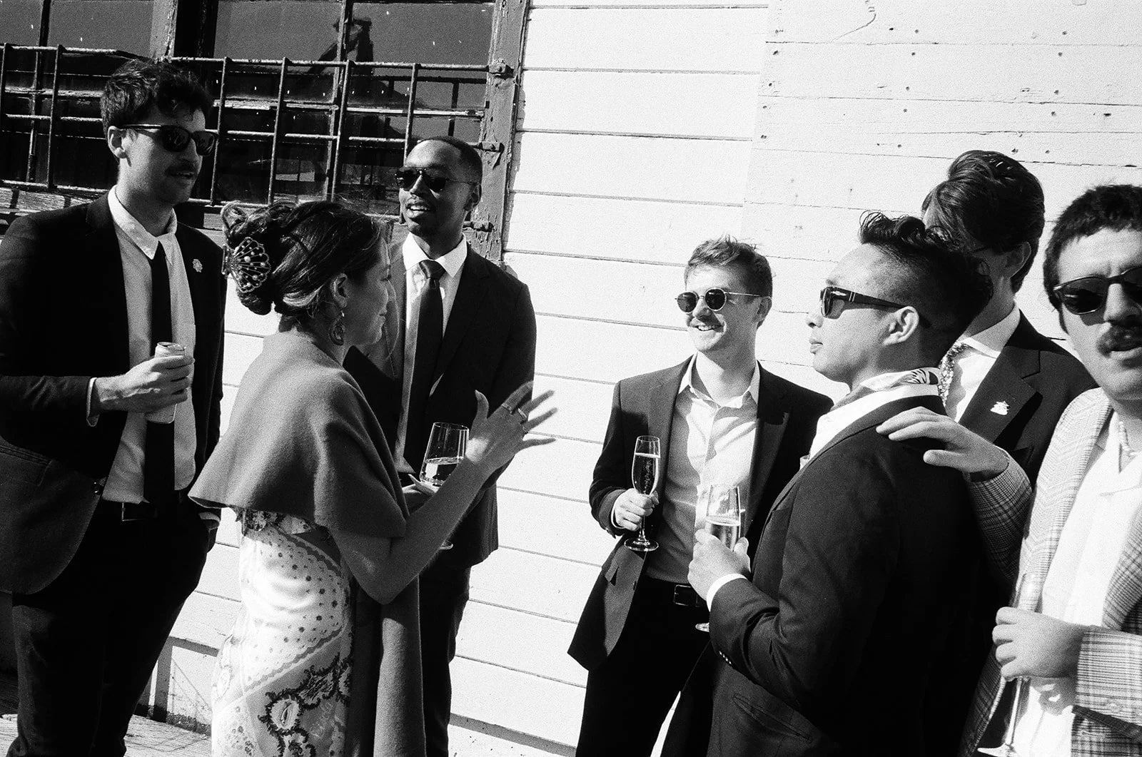 A group of seven people in formal attire, smiling and holding glasses of champagne, standing outdoors against a white wooden wall.