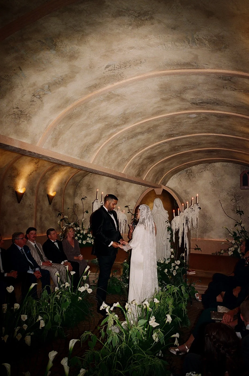 A wedding ceremony inside a chapel with an arched ceiling, featuring a couple exchanging vows in front of a religious statue, surrounded by guests seated on either side, and decorated with candles, flowers, and white drapery.
