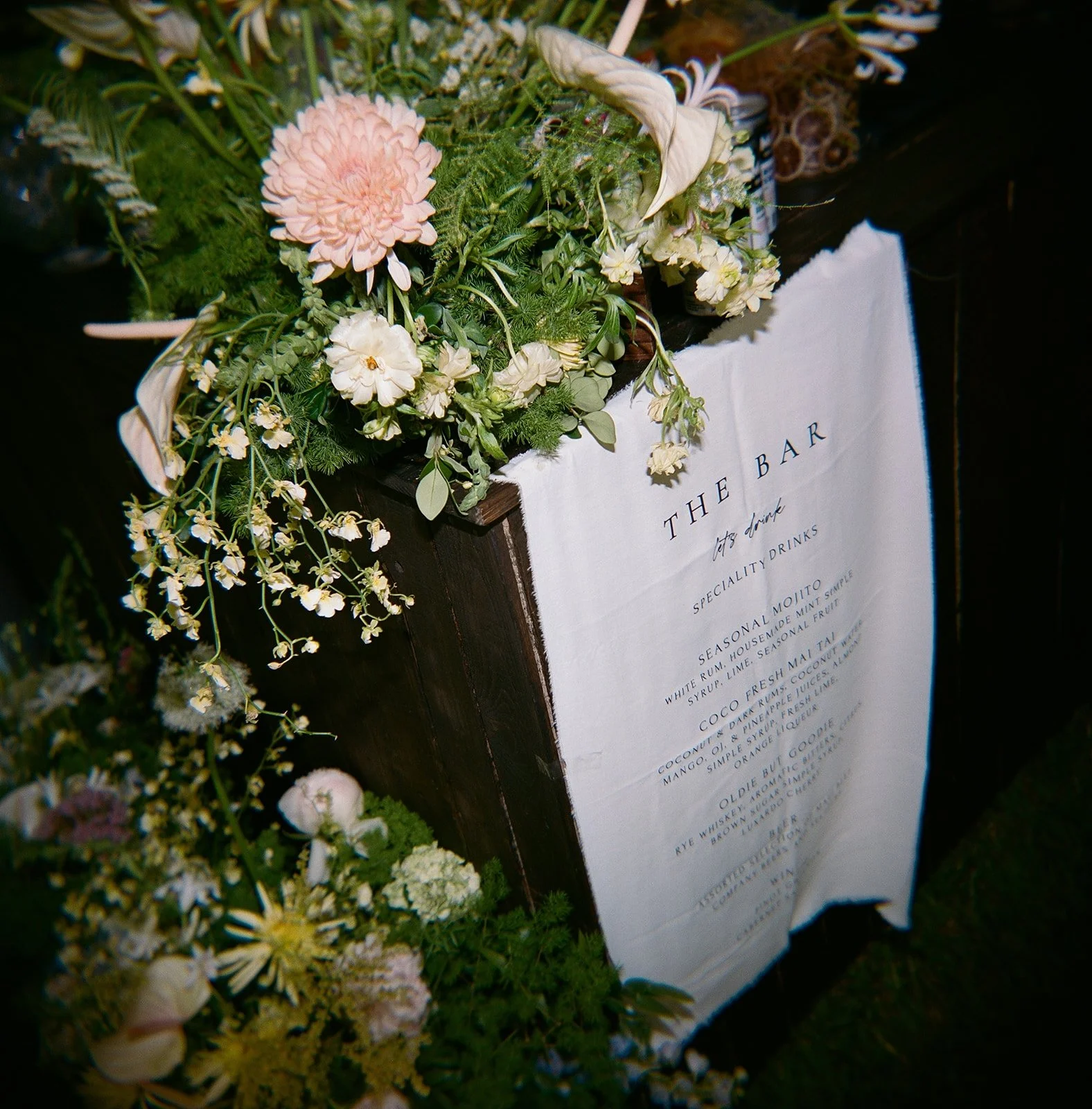 A wooden sign with a white cloth menu titled 'The Bar' decorated with white and pink flowers, including dahlias, roses, and greenery.