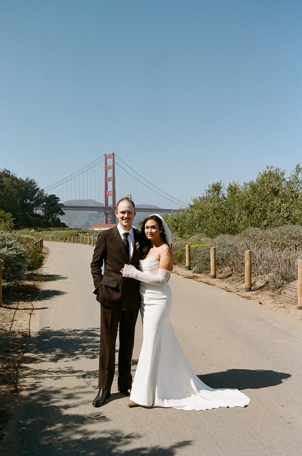 A bride and groom are standing on a path with the Golden Gate Bridge in the background, during a wedding photoshoot.