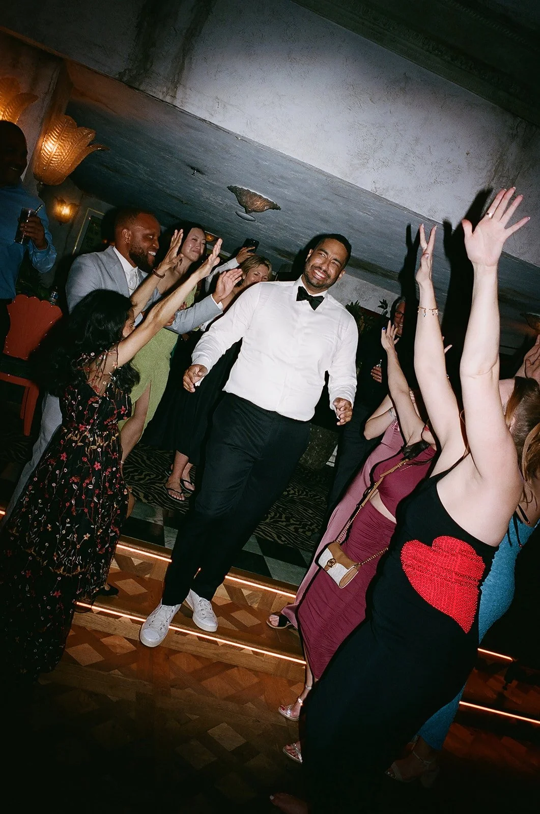 People dancing and celebrating at a party or event, man in a white shirt with a bow tie is smiling, others reaching out and dancing, festive atmosphere.