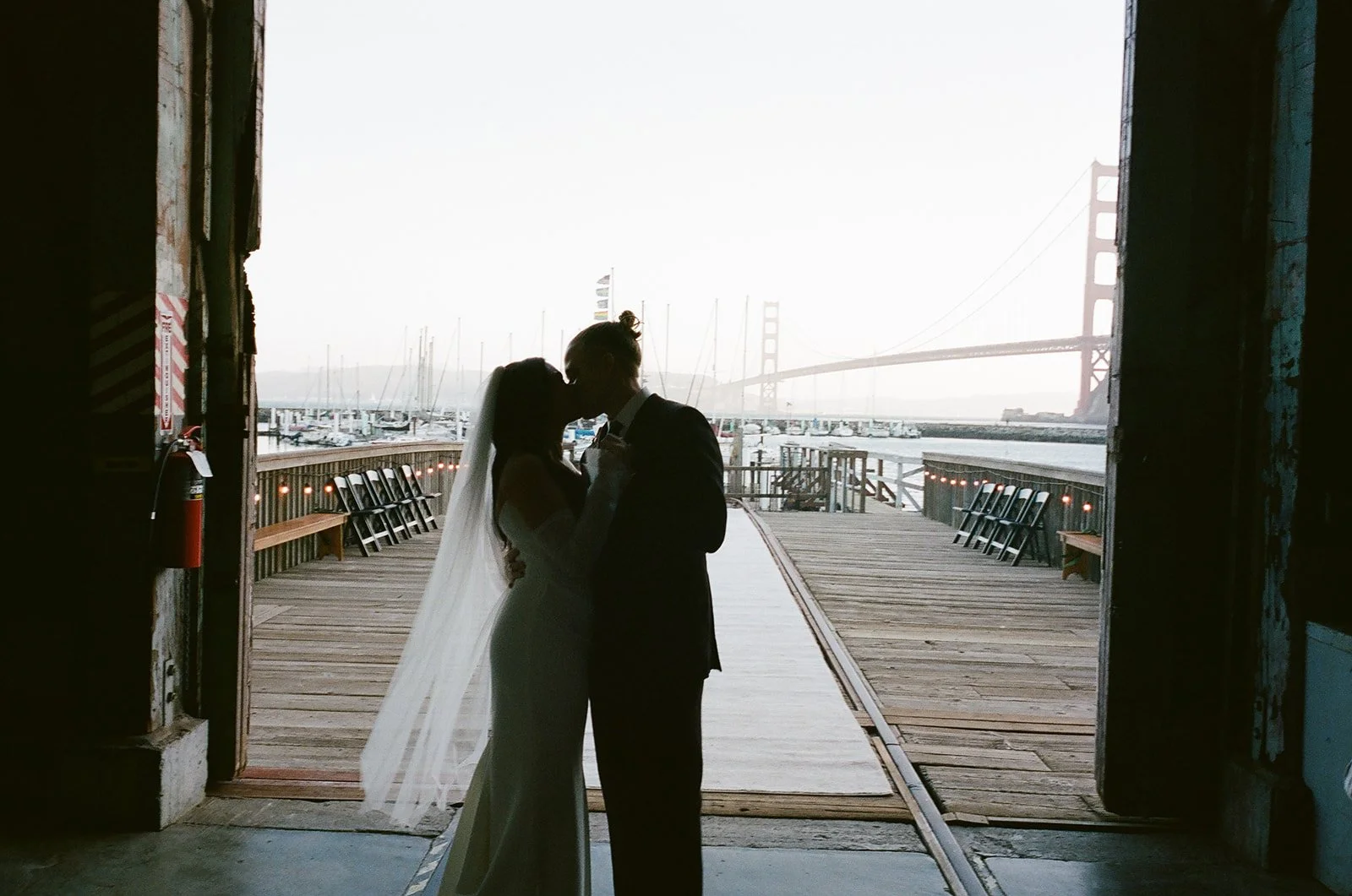 Silhouetted couple, in wedding attire, sharing a kiss at a dock with boats and the Golden Gate Bridge in the background.
