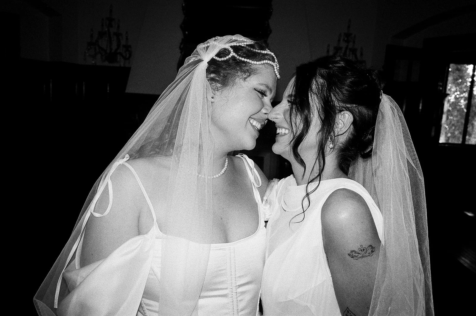Two women smiling and touching foreheads, dressed in wedding attire with veils, in an indoor setting.