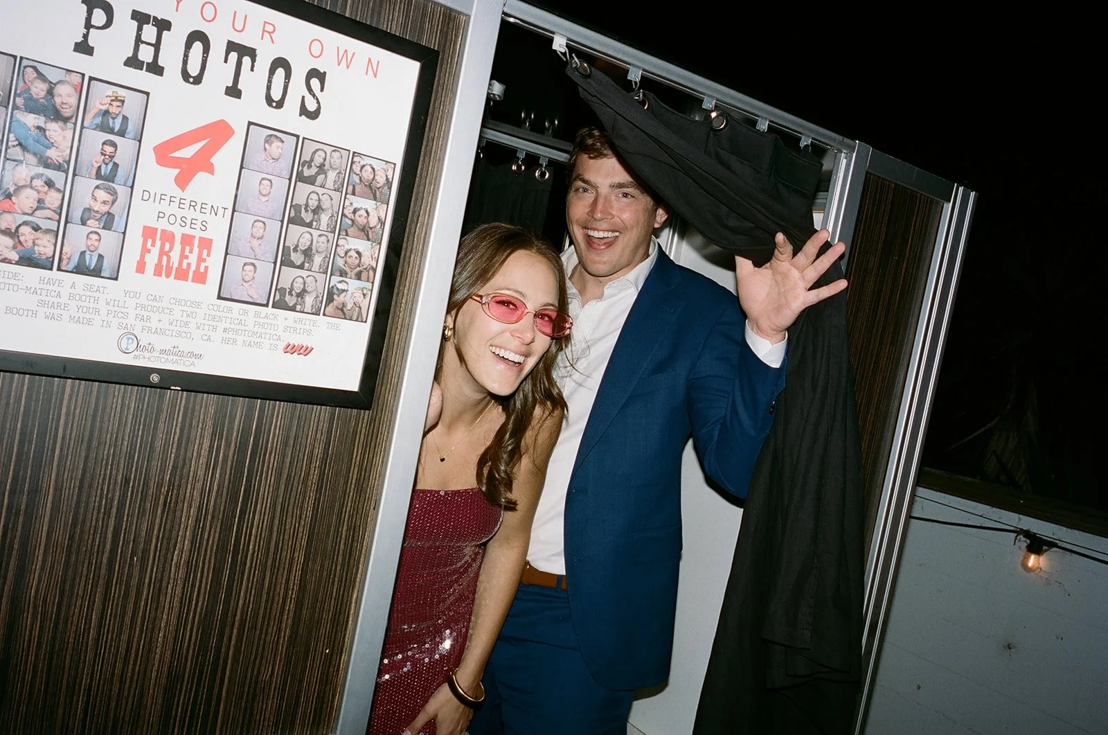 Two people in a photo booth, woman wearing a red dress and pink glasses, man in a blue suit holding a black curtain. They are smiling and posing for the camera.