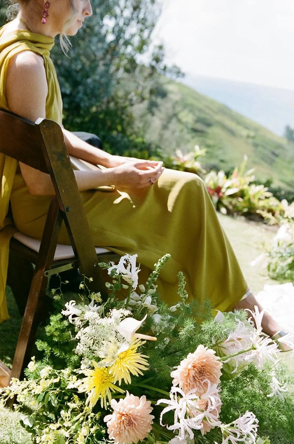 A woman sitting on a wooden chair outside, wearing a yellow dress and a matching scarf, with a large bouquet of pink and white flowers in front of her, overlooking a green hillside.
