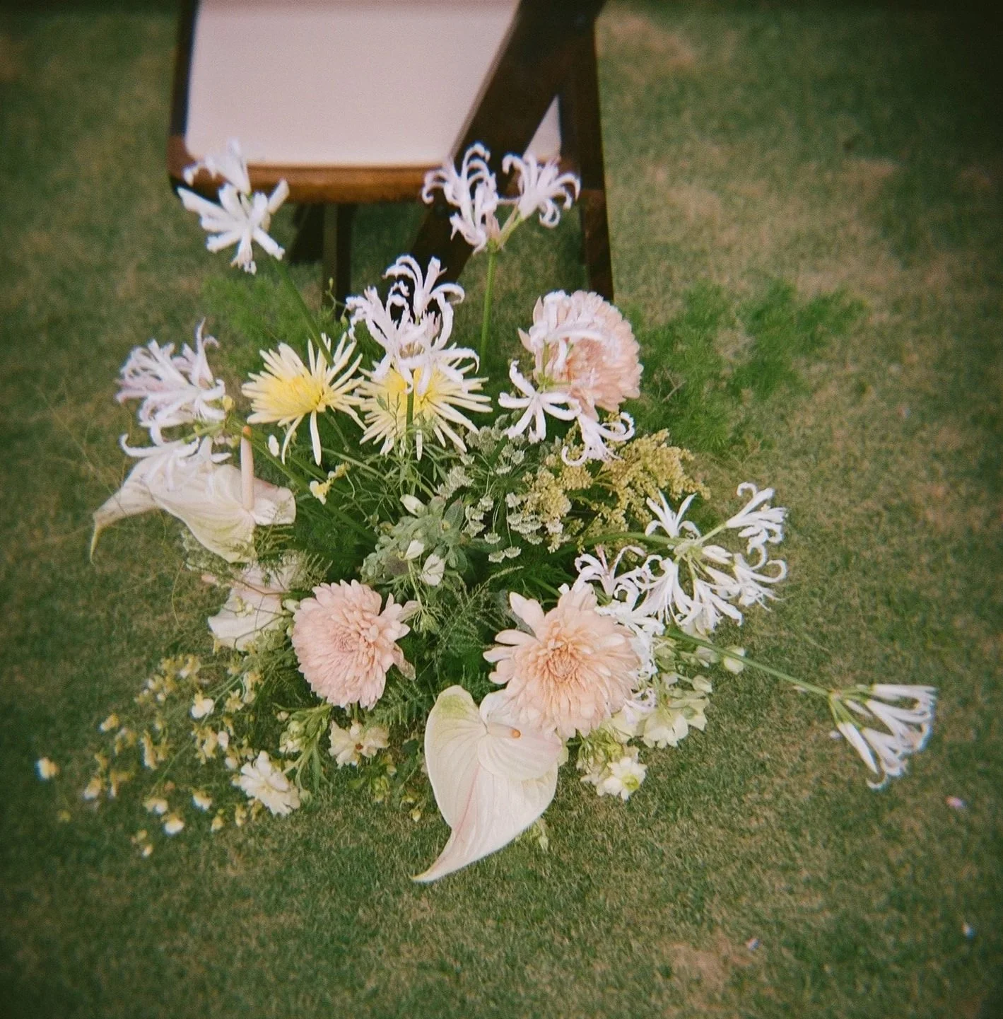 A colorful bouquet of mixed flowers including pink dahlias, white calla lilies, and spider lilies arranged on a grassy surface with a small table in the background.