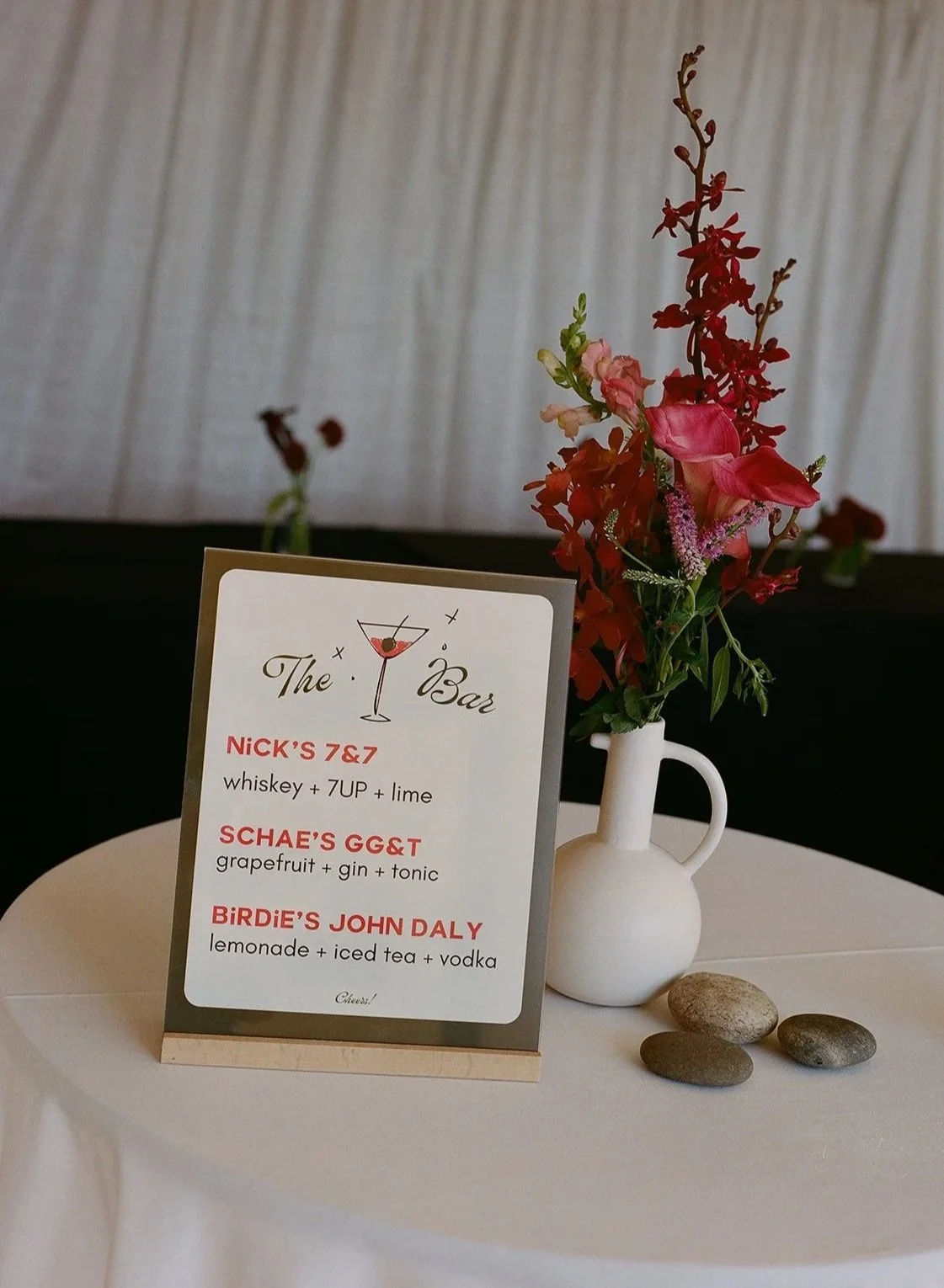 A table with a menu card and a white vase holding colorful flowers, and three small stones on a white tablecloth.