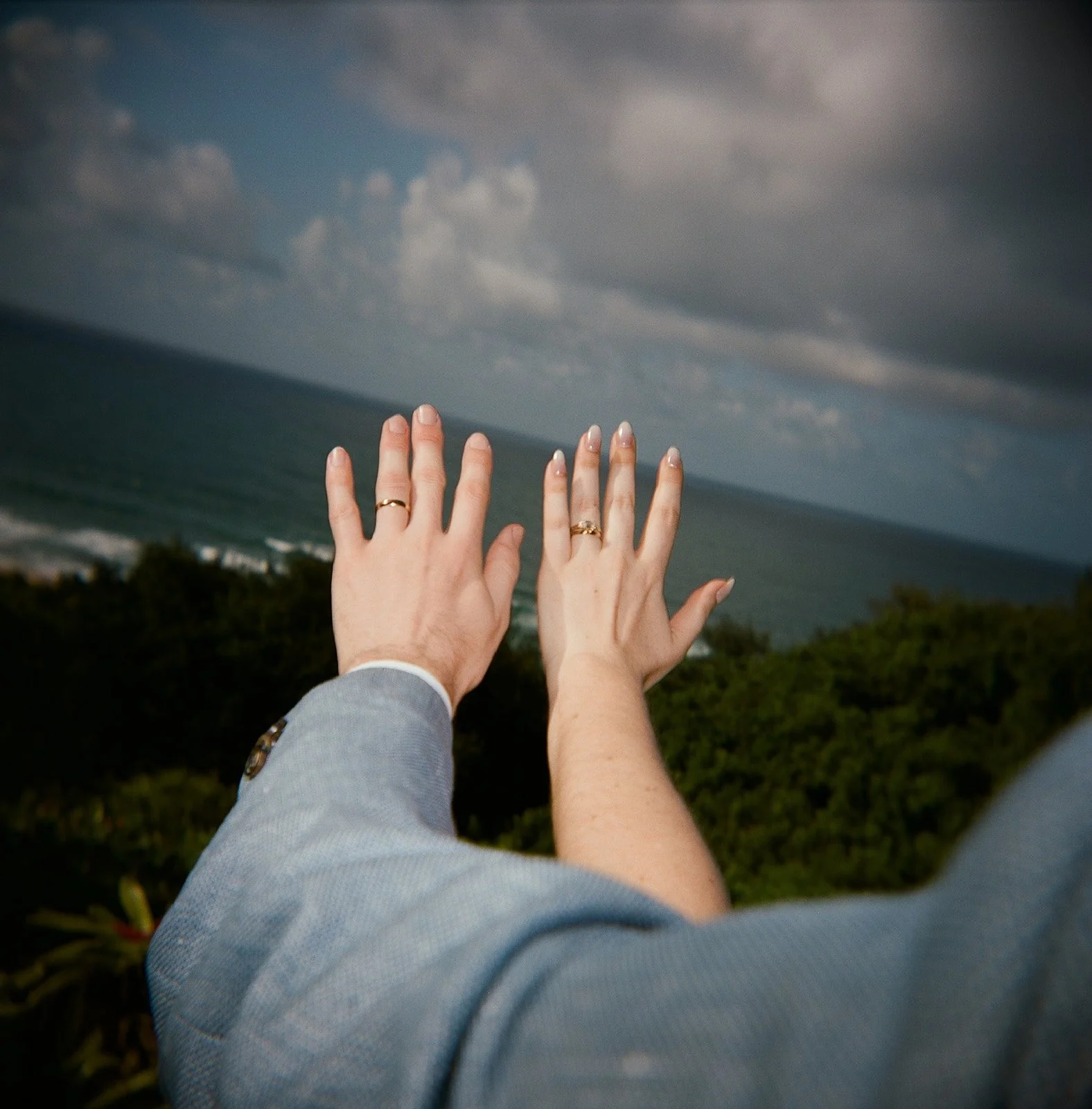 Two hands with wedding rings reaching out towards the sky over an ocean view with cloudy sky and green foliage below.