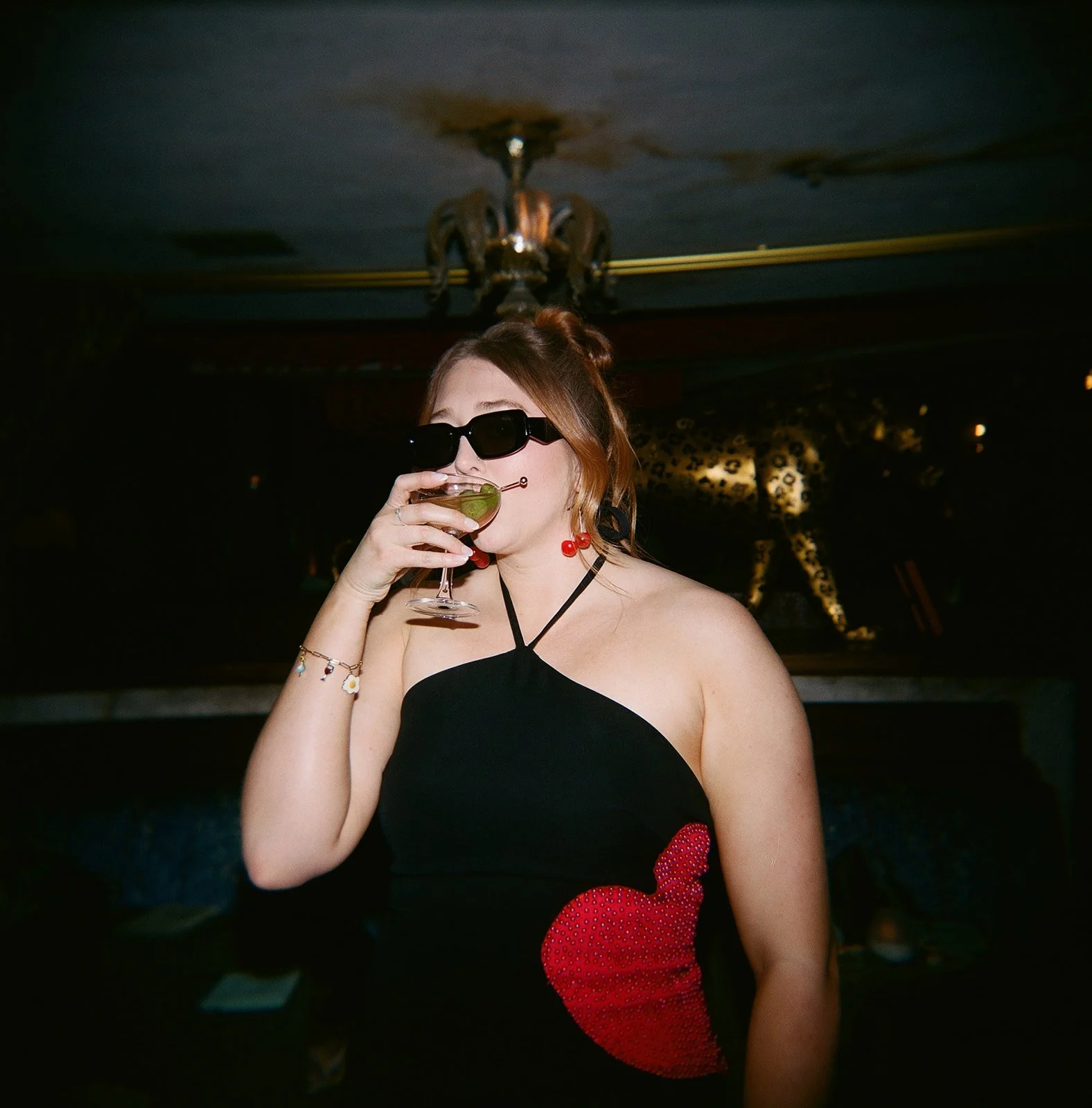 A woman wearing black sunglasses and a black dress with a red heart design is drinking a cocktail with a lime wedge in a dimly lit room.