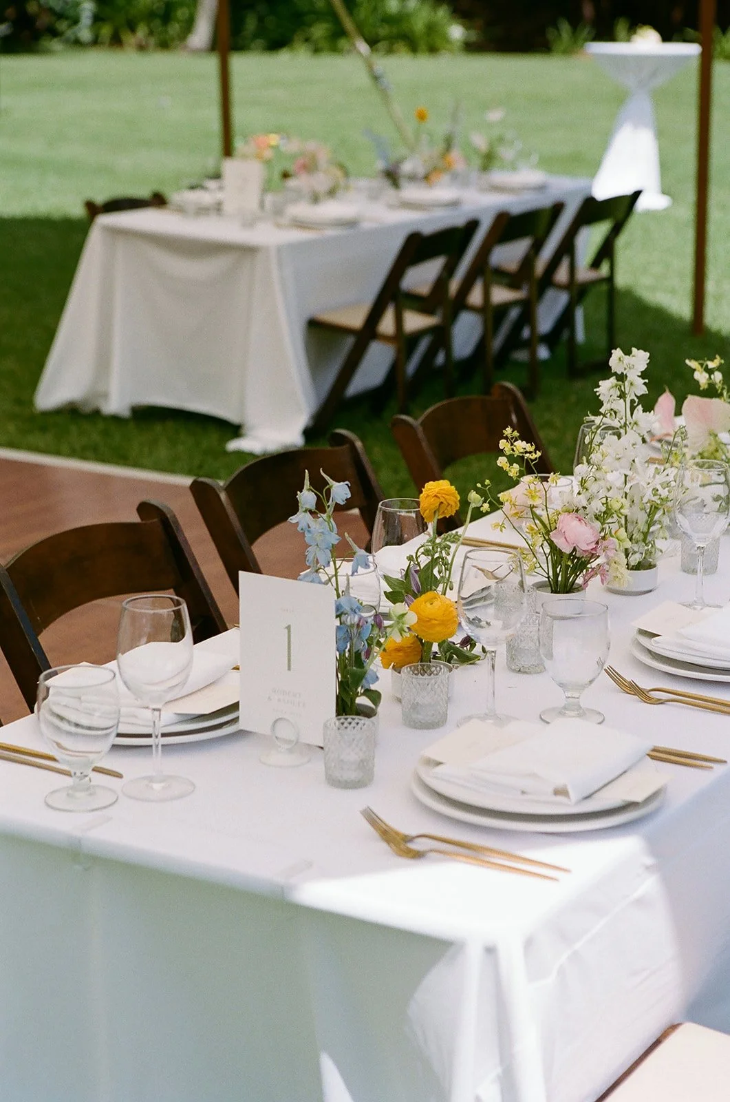 Decorated outdoor dining table with floral centerpieces, glassware, gold utensils, and numbered table card in a garden setting.