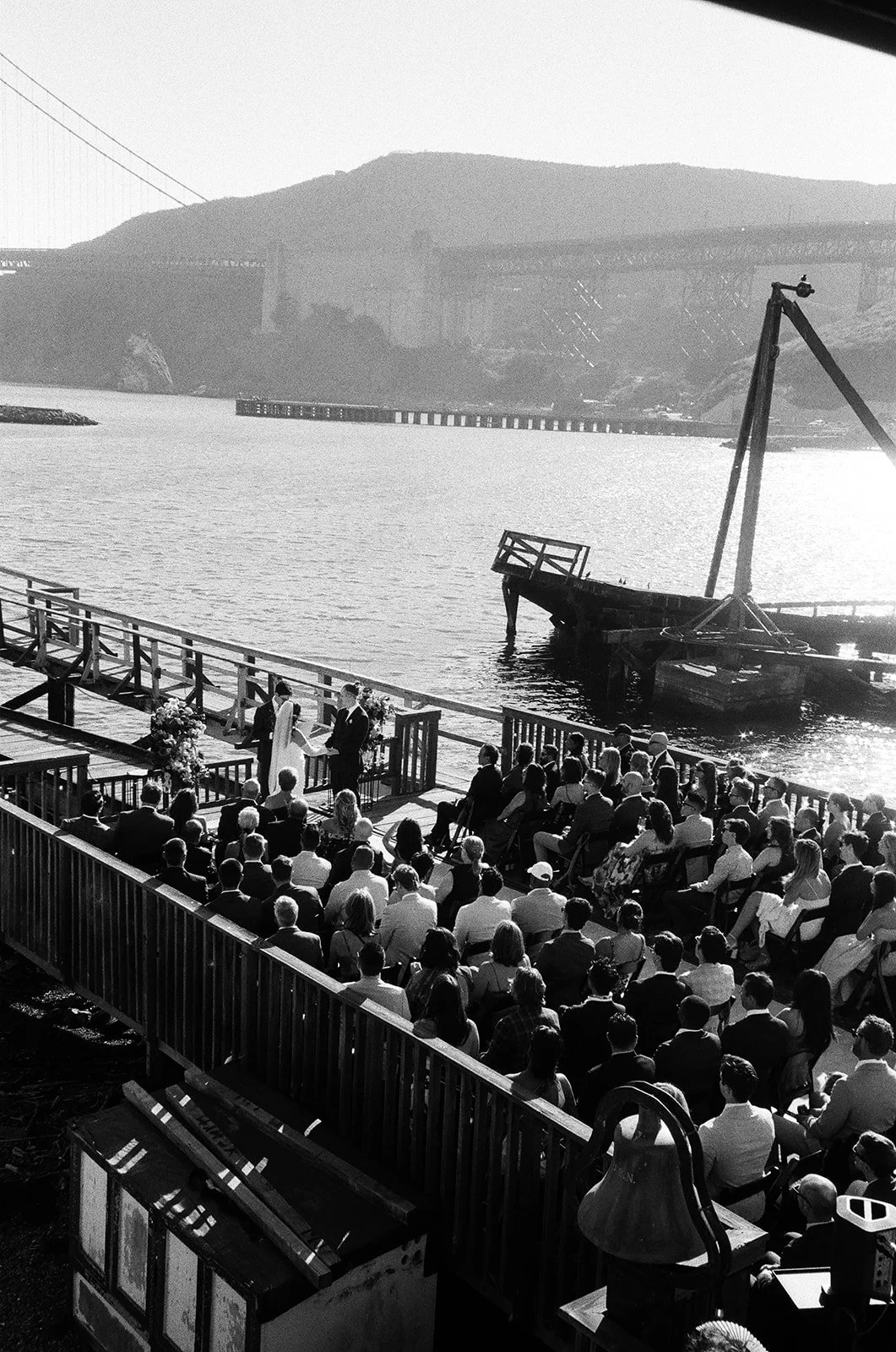 Black and white photo of a wedding ceremony taking place on a dock near the water, with a large audience sitting and standing, and a bridge and mountains in the background.