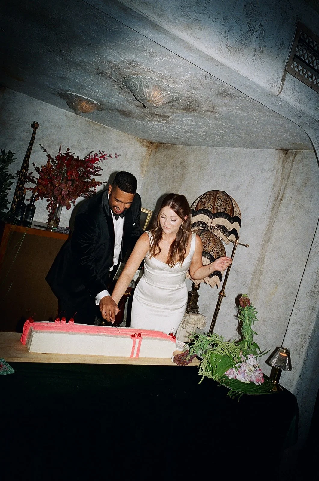 A couple in formal attire cutting a wedding cake at a celebration. The man is wearing a black tuxedo, and the woman is dressed in a white wedding gown. The scene is decorated with flowers, vintage lamps, and a textured wall background.