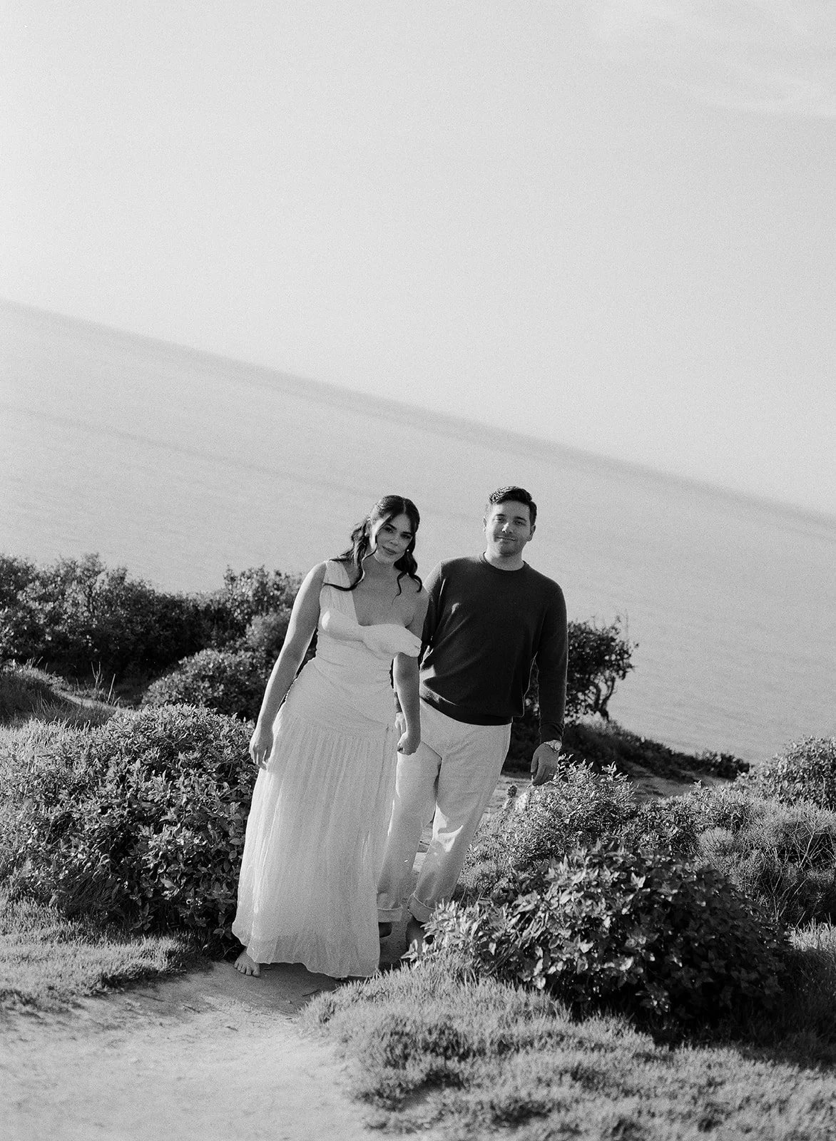 A black-and-white photo of a couple walking outdoors on a path in a grassy area with bushes, with the ocean visible in the background.