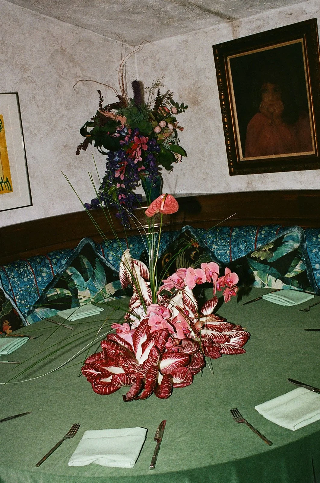 A table with a centerpiece consisting of pink and red plants, surrounded by napkins and silverware, in a dining room with floral and artwork decor.