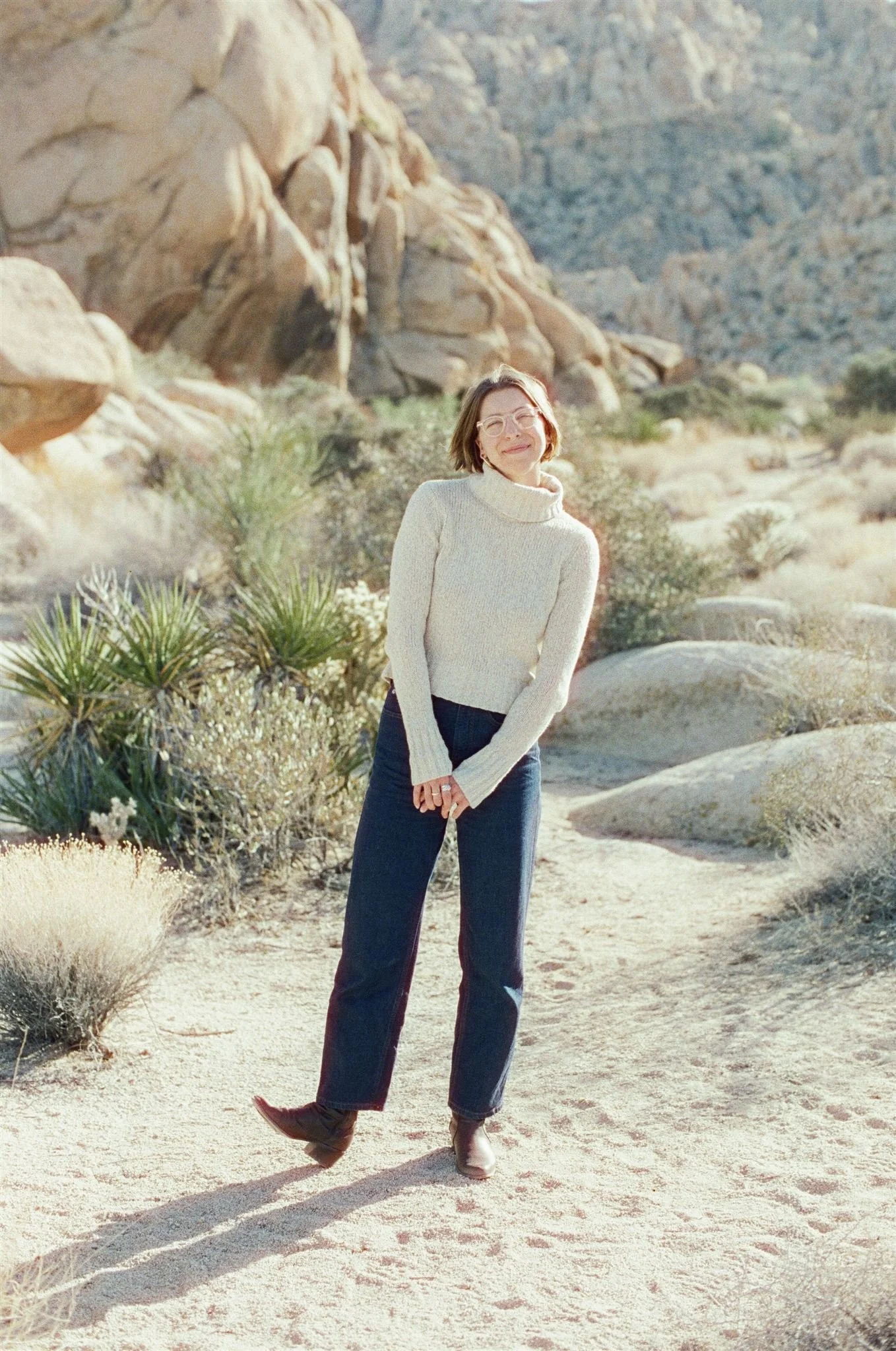 A woman stands outdoors in a desert landscape with rocks and sparse vegetation, smiling and wearing a cream-colored turtleneck sweater, dark jeans, and boots.
