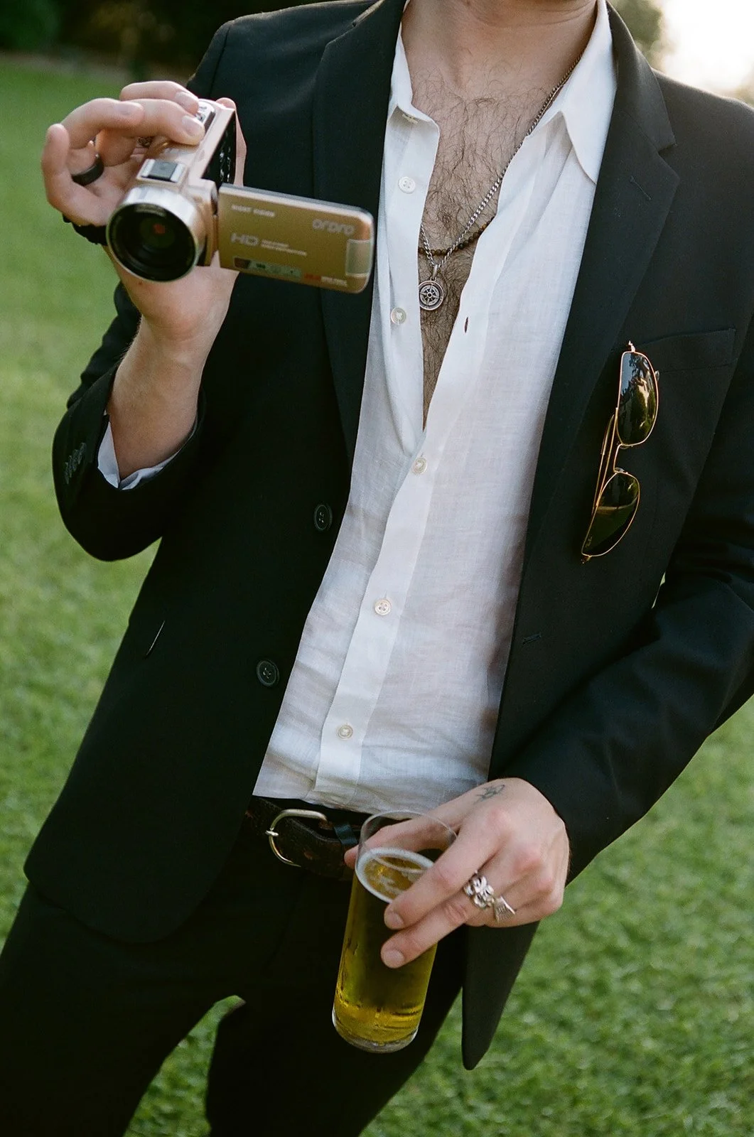 A man in a black suit jacket and white shirt holding a digital camcorder and a pint glass of beer, with sunglasses hanging from his pocket, standing outdoors on grass.