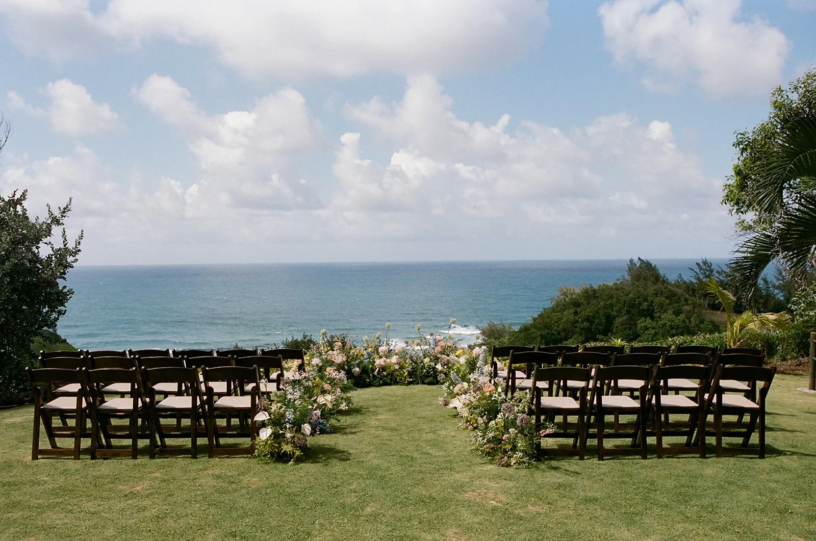 Outdoor wedding setup with chairs, floral decorations, overlooking the ocean under a partly cloudy sky.