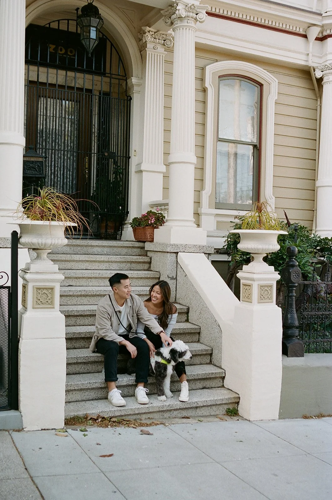 A young couple sitting on the steps of a Victorian-style house with a dog, smiling and interacting with each other.
