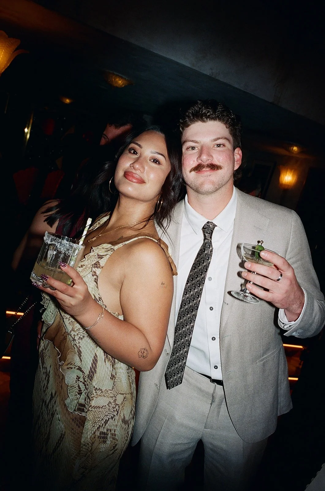 A young woman and a young man enjoying drinks at a social event, holding cocktails and smiling at the camera.