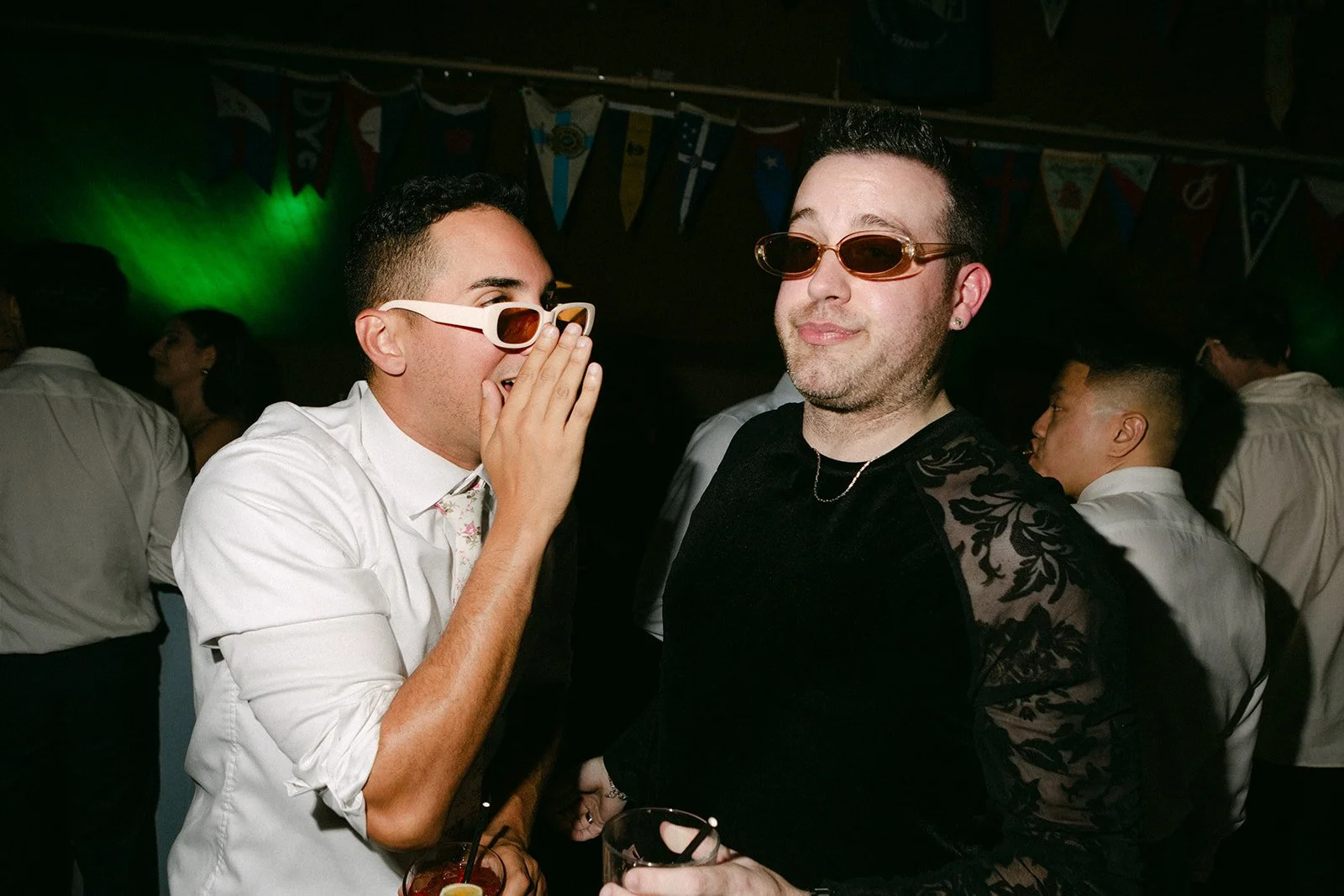 Two men wearing sunglasses at a party, one whispering to the other. Background shows other people and colorful banners hanging from the ceiling.