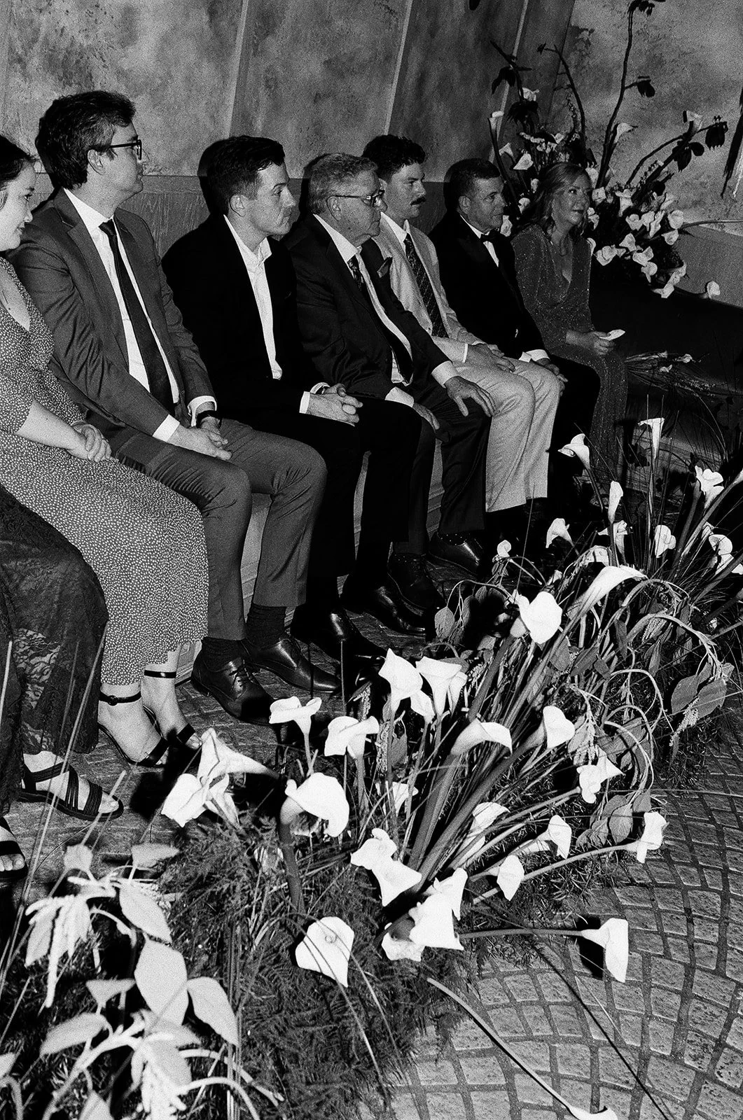A group of seven people seated on a bench with flowers at their feet, attending an indoor event.