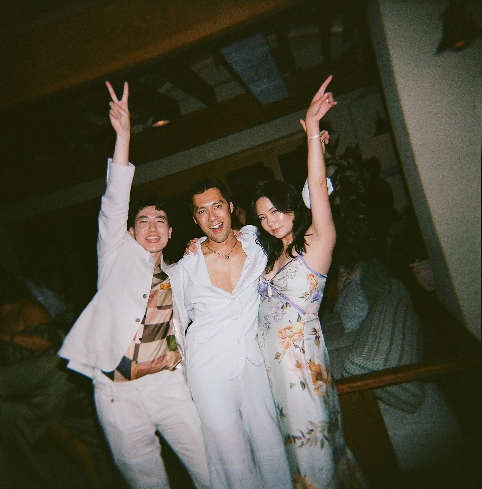 Three friends celebrating at a party, two men and one woman, smiling and making peace signs with their hands.
