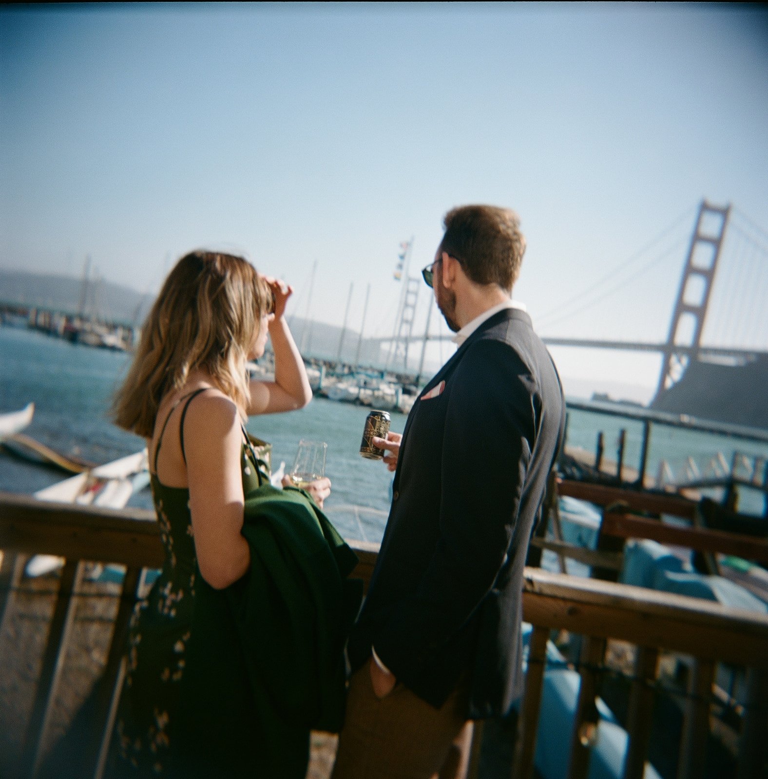 A man and woman standing outdoors near a body of water with boats and a bridge in the background, engaging in conversation while holding drinks.