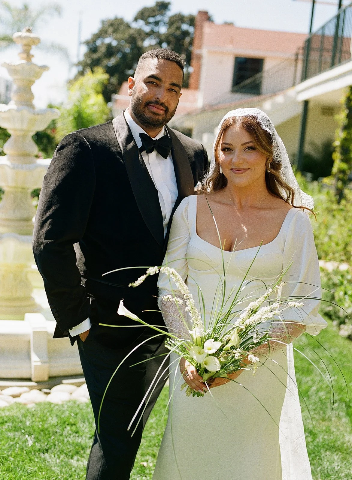 A bride and groom posing outdoors in a garden setting on their wedding day. The groom is in a black tuxedo with a bow tie, and the bride is wearing a cream-colored wedding dress and holding a bouquet of white flowers.