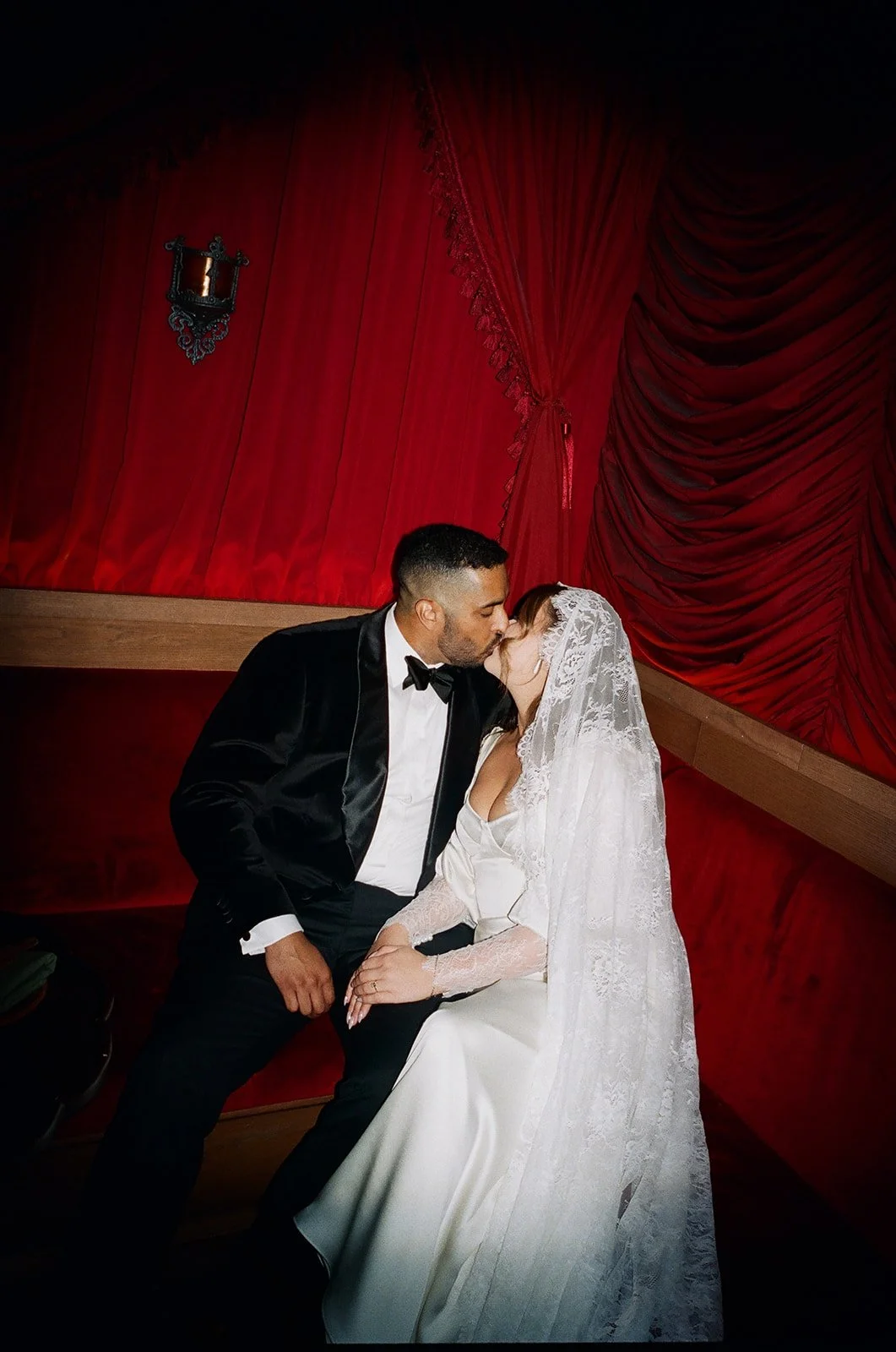 A bride and groom sharing a kiss in a dimly lit room with red velvet curtains and a wall sconce.