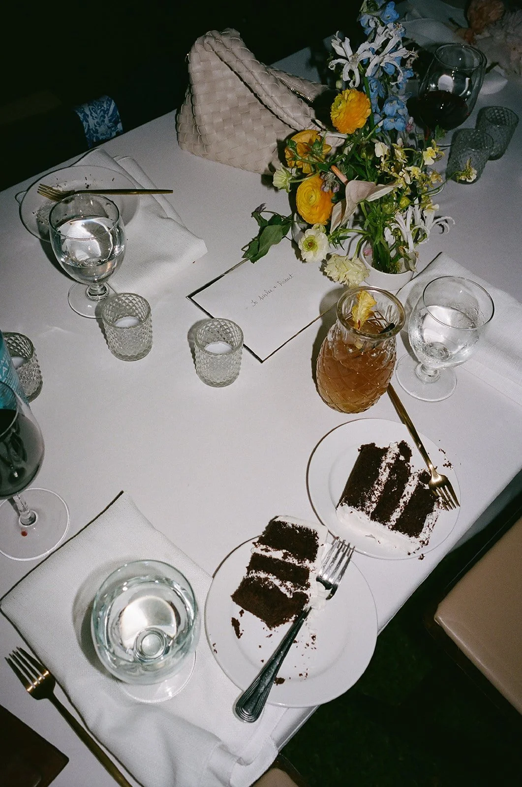 A dining table set for a celebration with a flower arrangement, various glasses filled with water and drinks, and a plate with partially eaten chocolate cake.