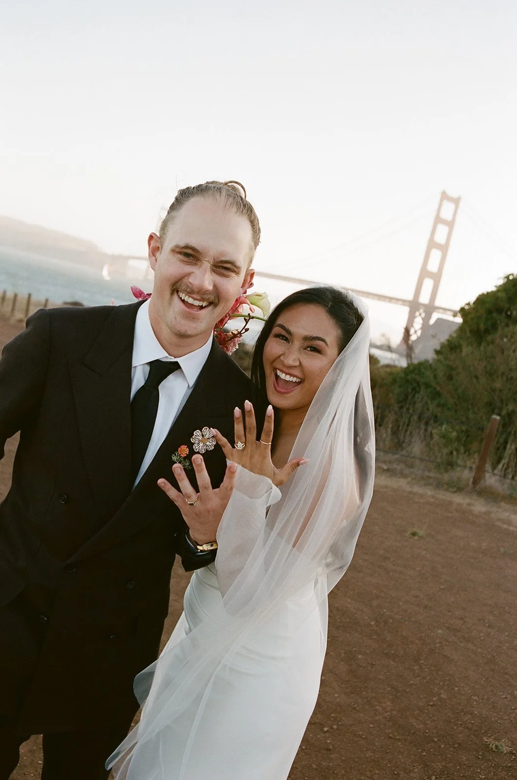 A happy couple on their wedding day, displaying their rings, with the Golden Gate Bridge in the background.