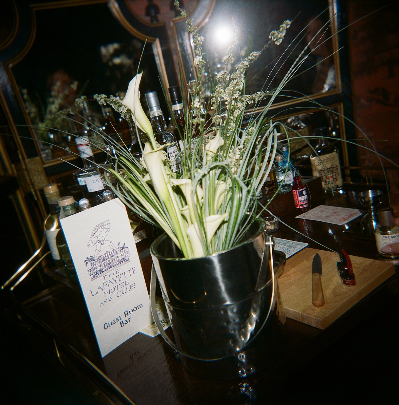 A floral arrangement of white calla lilies and greenery in a metallic bucket on a bar counter at The Lafayette Hotel and Club, with a guest room bar sign nearby.
