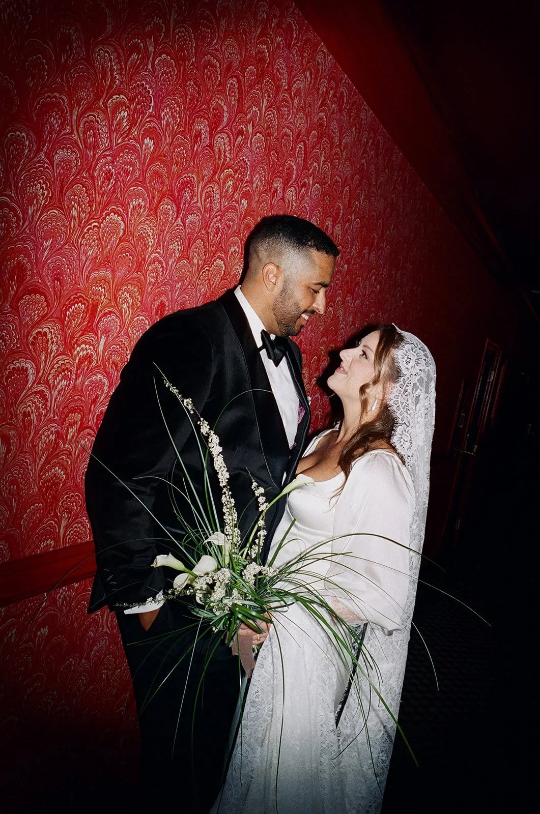 A bride and groom share a moment of intimacy, standing close against a red patterned wall. The bride wears a white wedding dress and veil, holding a white floral bouquet. The groom is dressed in a black tuxedo with a bowtie.