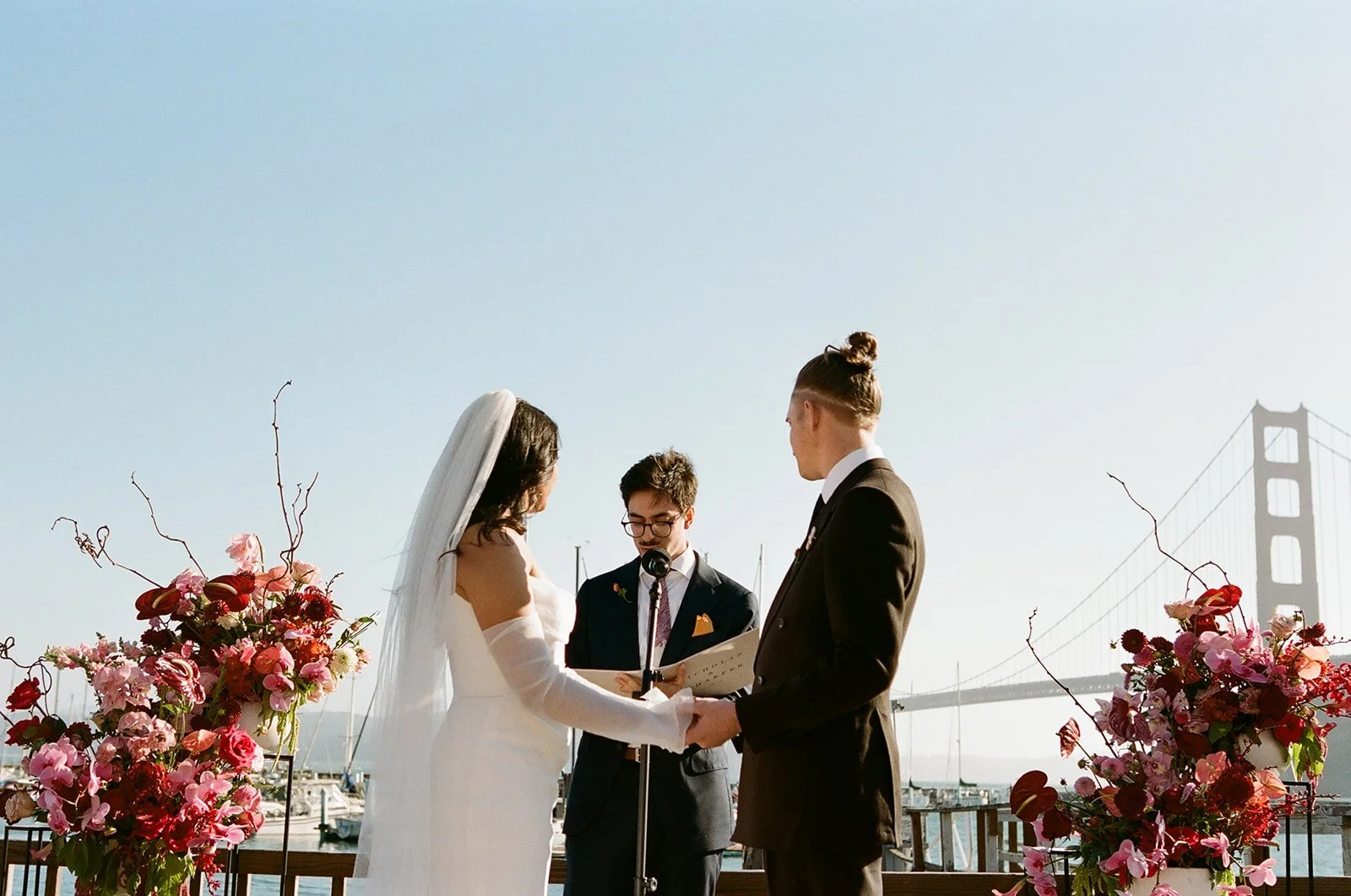 A couple getting married outdoors by the water with the Golden Gate Bridge in the background. The bride is in a white wedding dress and veil, and the groom is in a black suit. They are holding hands and facing each other during the ceremony, with a p