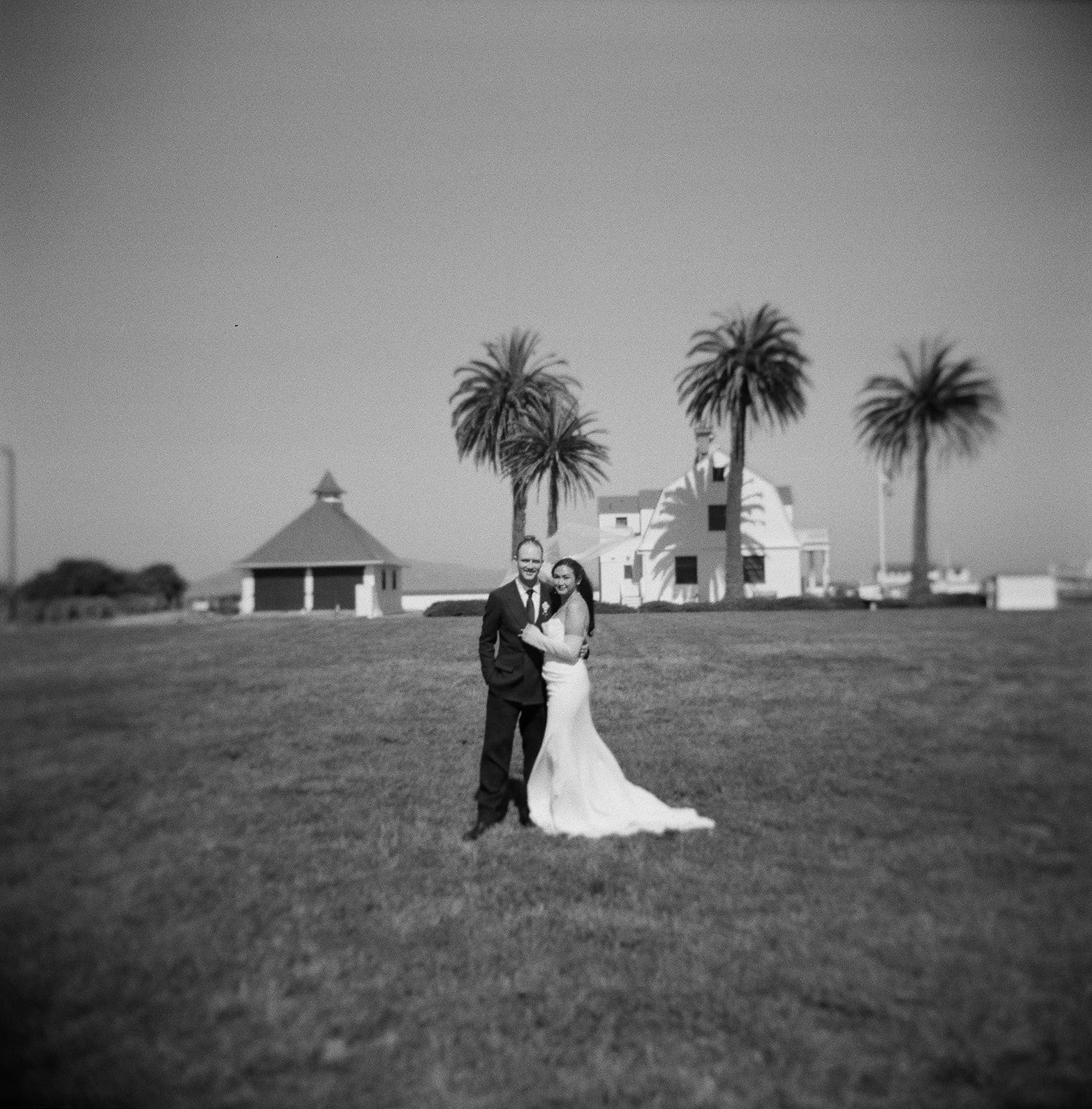 A black and white photo of a newlywed couple standing on a grassy lawn with palm trees and a white house in the background.