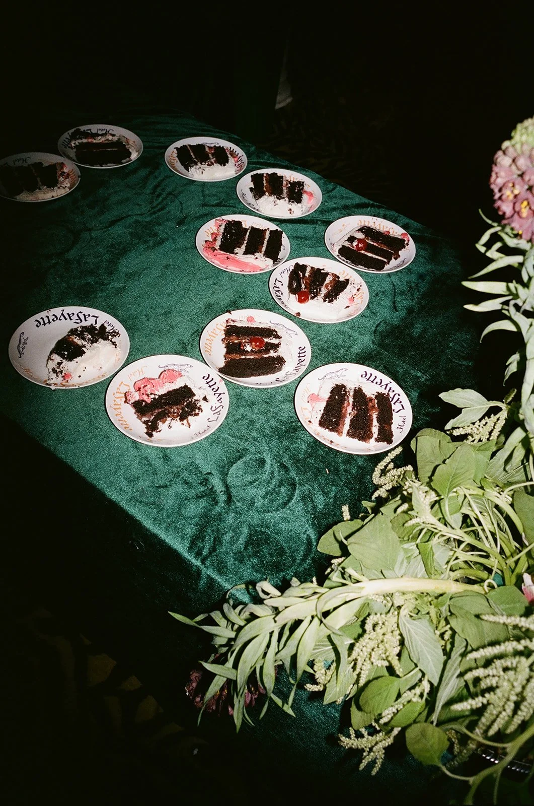 Several plates with slices of chocolate cake on a green tablecloth, some with cherries on top, and a floral arrangement on the right side.