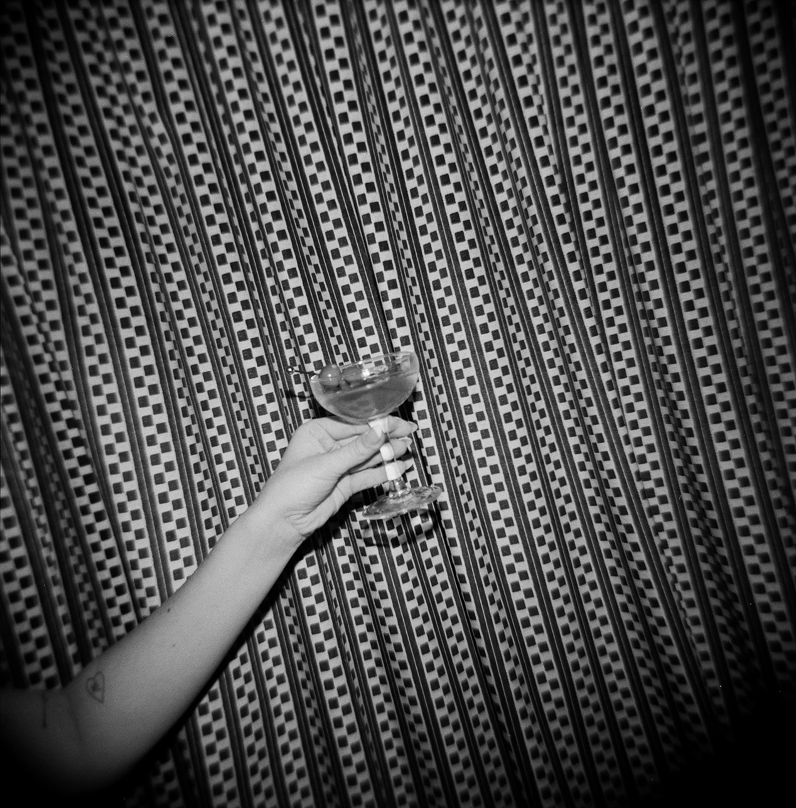 A person's hand with a small tattoo on their wrist holding a glass of dark beverage against a patterned fabric background.