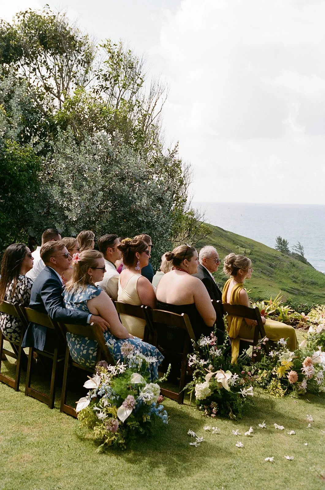 People sitting outdoors at a wedding ceremony, with a scenic view of the ocean and greenery in the background, decorated with flowers.